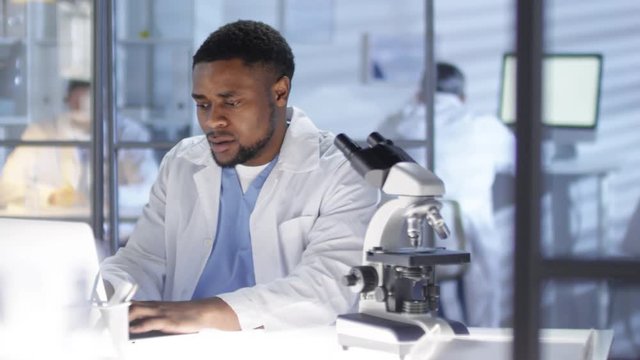 Panning waist-up shot of African American research scientist, wearing medical uniform, sitting at desk in lab and typing on laptop, with microscope by side, then looking at camera and smiling happily