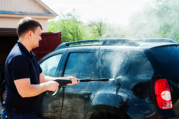 A man washes a black car from a hose with high-pressure water near the garage