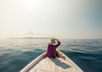 Young woman sitting on bow of boat in the middle of the sea. Happy girl with hat raising hands enjoying freedom travel.