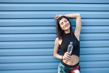 Beautiful young fitness brunette girl in the top and the leggings with water bottle after a hard workout, standing near blue vinyl plastic panels texture wall. Sport and healthy lifestyle