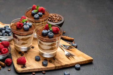 Classic tiramisu dessert with blueberries and raspberries in a glass on wooden cutting board on dark concrete background