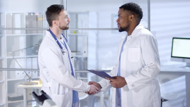 Thigh-up shot of friendly Caucasian and African American doctors, dressed in blue scrubs and lab coats, meeting in hospital staff room, shaking hands, and discussing work issues