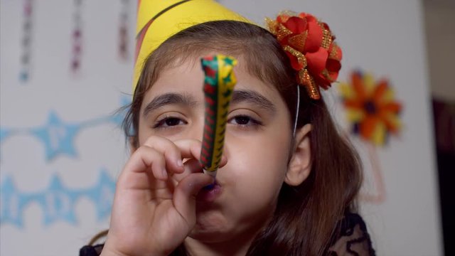 Cute Indian girl blowing blow out whistle at a birthday party - Fun at home. Closeup shot of a girl playing at a birthday bash