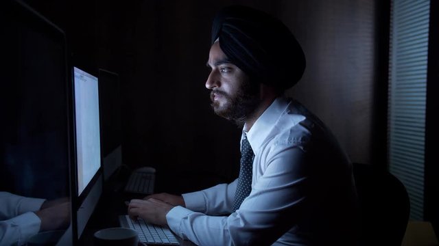 Tired exhausted employee drinking coffee while working late night in the office. HD stock video of a young Indian man wearing a turban and formal clothes working and having coffee at work