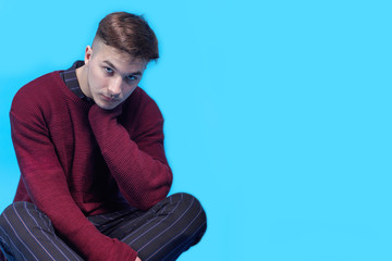 Young handsome man sitting on the floor on blue background.  Fashion hairstyle, expressive harsh look to the camera. Slight beard, burgundy sweater. Indoors, isolated, big copy space.