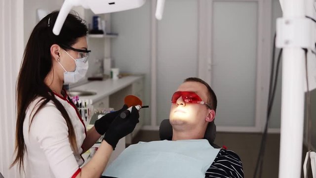 Female doctor and male patient in protective orange glasses in the modern dental ocabinet. Dentist is doing polymerisation of fillings with a orange protective screen