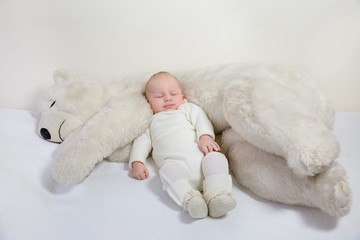 View of a young infant sleeping with a huge white teddy bear on a bed.