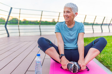 Relaxed athletic mature woman sitting on fitness mat outdoors. Senior Woman Resting After Exercises. Woman on a yoga mat to relax outdoor. Senior lady prefers healthy lifestyle