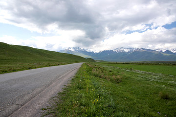 Fototapeta na wymiar Country road among green fields and hills against the backdrop of mountains with snow-capped peaks