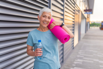 Portrait of confident mature woman with exercise mat smiling. Woman holding rolled up exercise mat. Happy mature woman with a yoga mat. Fit Senior Woman With Exercise Mat
