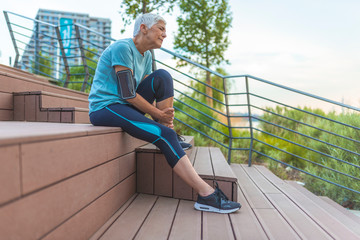 Senior woman In Sportswear Sitting And Touching Painful Ankle Injured While Training Outdoors. Sports Injury. High Resolution. Sport runner woman cramps in leg and feel pain