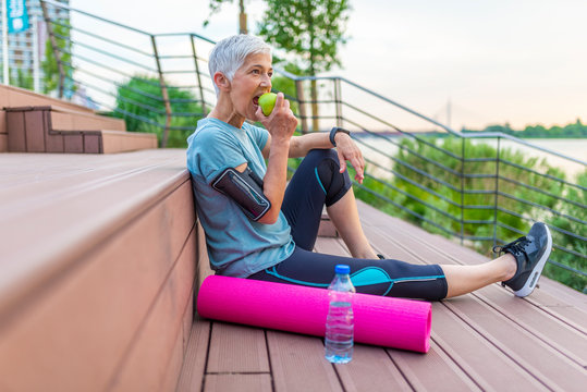 Sport mature woman sitting and resting after workout or exercise and eating apple on floor. Relax concept. Strength training and Body build up theme. Beautiful sporty woman eating apple