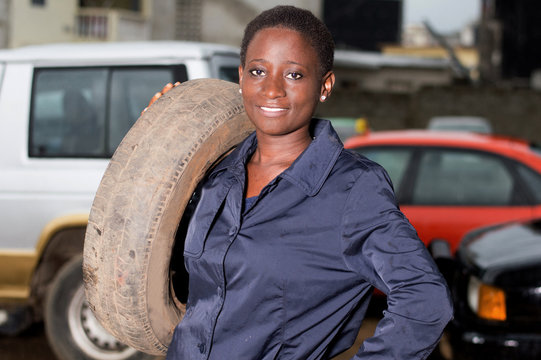 Young mechanic standing and smiling has a tire on her shoulder in her studio.
