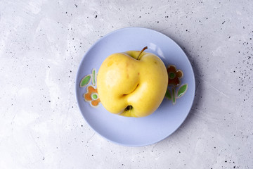 One ugly yellow apple on lilac plate on grey concrete background. 