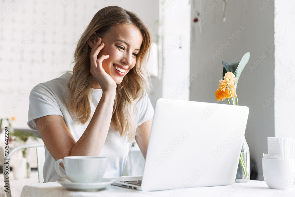 Poster young smiling pretty blonde woman student sitting in cafe using laptop computer drinking coffee.