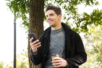 Handsome happy man in casual clothing walking outdoors in green park using mobile phone drinking...