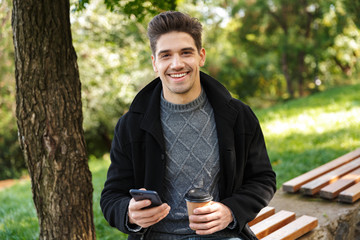 Handsome cheerful young happy man in casual clothing walking outdoors in green park using mobile phone drinking coffee.