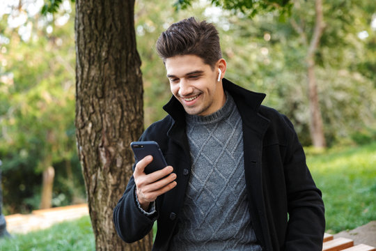 Smiling Handsome Young Cheerful Man In Casual Clothing Walking Outdoors In Green Park Using Mobile Phone Listening Music With Earphones.