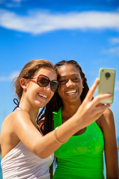 2 smiling girls taking a selfie against blue sky background.