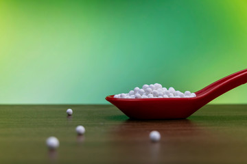 Homeopathic pills in red spoon and some globules scattered on wooden surface with green mix yellow background