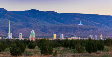 Wednesday, 26 June 2019; Ashgabat, Turkmenistan; Beautiful cityscape, panorama view on the white city at blue mountains with iconic landmarks of the capital of Central Asian state few minutes sunset. - obrazy, fototapety, plakaty
