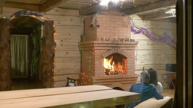 Couple is sitting by brightly burning fireplace in wooden house, view of room outside through window, stock video. Red brick fireplace with hot flame, place for family holidays and romantic date