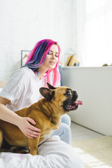 girl with colorful hair petting bulldog looking away while sitting on bed