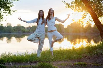 two yogin  happy girls practicing yoga in white clothes  in the rays of the sunset sun on a lush green meadow at the lake