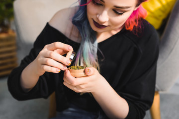beautiful girl with colorful hair holding and looking at herb grinder while sitting on floor