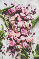 Flat-lay of sweet pink macaron cookies, lilac marshmallows and spring flowers over wooden background, top view, vertical composition. Food texture, background and wallpaper