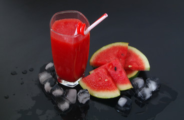 Glass of watermelon smoothie with ice cubes and watermelon slices on black table 