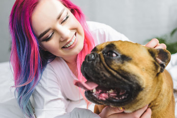 selective focus of happy girl with colorful hair laying with french bulldog in bed