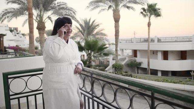 Smiling Caucasian senior woman talking on a smartphone on a hotel terrace in a white terry robe at a tropical resort. Against the backdrop of palm trees and the sea. The concept of using gadgets by