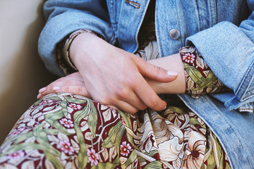 mid section of young woman wearing denim jacket over summer dress with floral pattern - close-up of female hands folded in her lap - Powered by Adobe