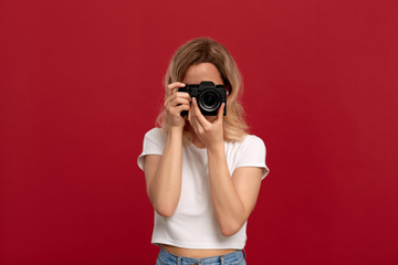 Portrait of a girl with curly blond hair dressed in a white t-shirt standing on a red background. Model takes photos with retro film camera.