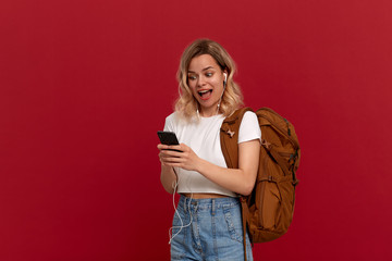 Fototapeta na wymiar Portrait of a girl with curly blond hair dressed in a white t-shirt standing on a red background. Excited model with orange backpack and white headset looks at the phone screen.