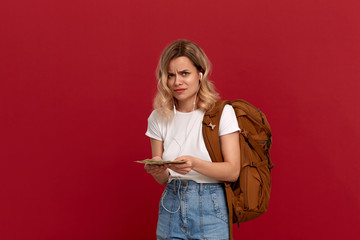 Portrait of a girl with curly blond hair, orange backpack and white headset dressed in a white t-shirt standing on a red background expressing puzzled emotion because she doesn't have enough money.