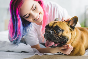 selective focus of girl with colorful hair petting and looking at cute french bulldog while laying in bed