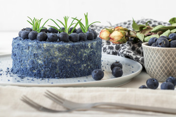 Table with Blue Blueberry Velvet cake. Cup of coffee with milk, small bowl with blueberries, scarf and dry flowers, roses.
