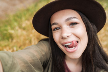 Photo closeup of cheerful woman wearing hat showing her tongue at camera while taking selfie photo...