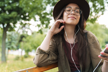 Photo of pleased woman wearing hat and eyeglasses using earphones and smartphone in green park