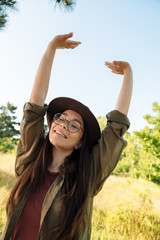 Photo of happy brunette woman wearing stylish hat and eyeglasses walking in green park on sunny day