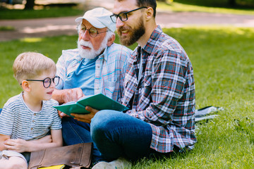 happy family of three generation - father, grandfather and cute blond son sitting on grass at park with books learn to read while getting ready for school.