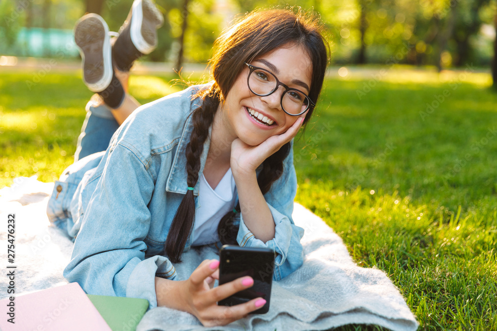 Canvas Prints cheerful smiling cute young student girl wearing eyeglasses outdoors in nature park using mobile pho