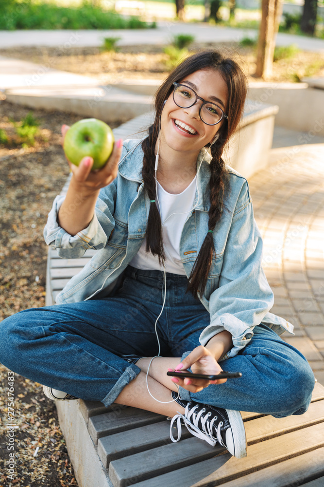 Sticker Positive happy cute young student girl wearing eyeglasses sitting on bench outdoors in nature park using mobile phone chatting.