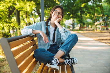Pleased happy cute young student girl wearing eyeglasses sitting on bench outdoors in nature park