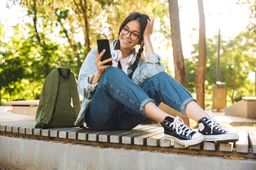 Positive happy cute young student girl wearing eyeglasses sitting on bench outdoors in nature park...