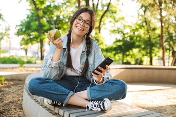 Positive happy cute young student girl wearing eyeglasses sitting on bench outdoors in nature park using mobile phone chatting.