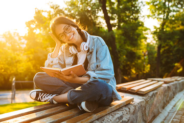 Happy cheerful smiling young student girl wearing eyeglasses sitting outdoors in nature park...