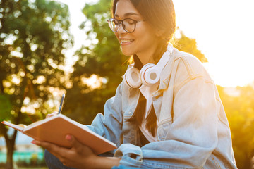 Happy young student girl wearing eyeglasses sitting outdoors in nature park listening music with...